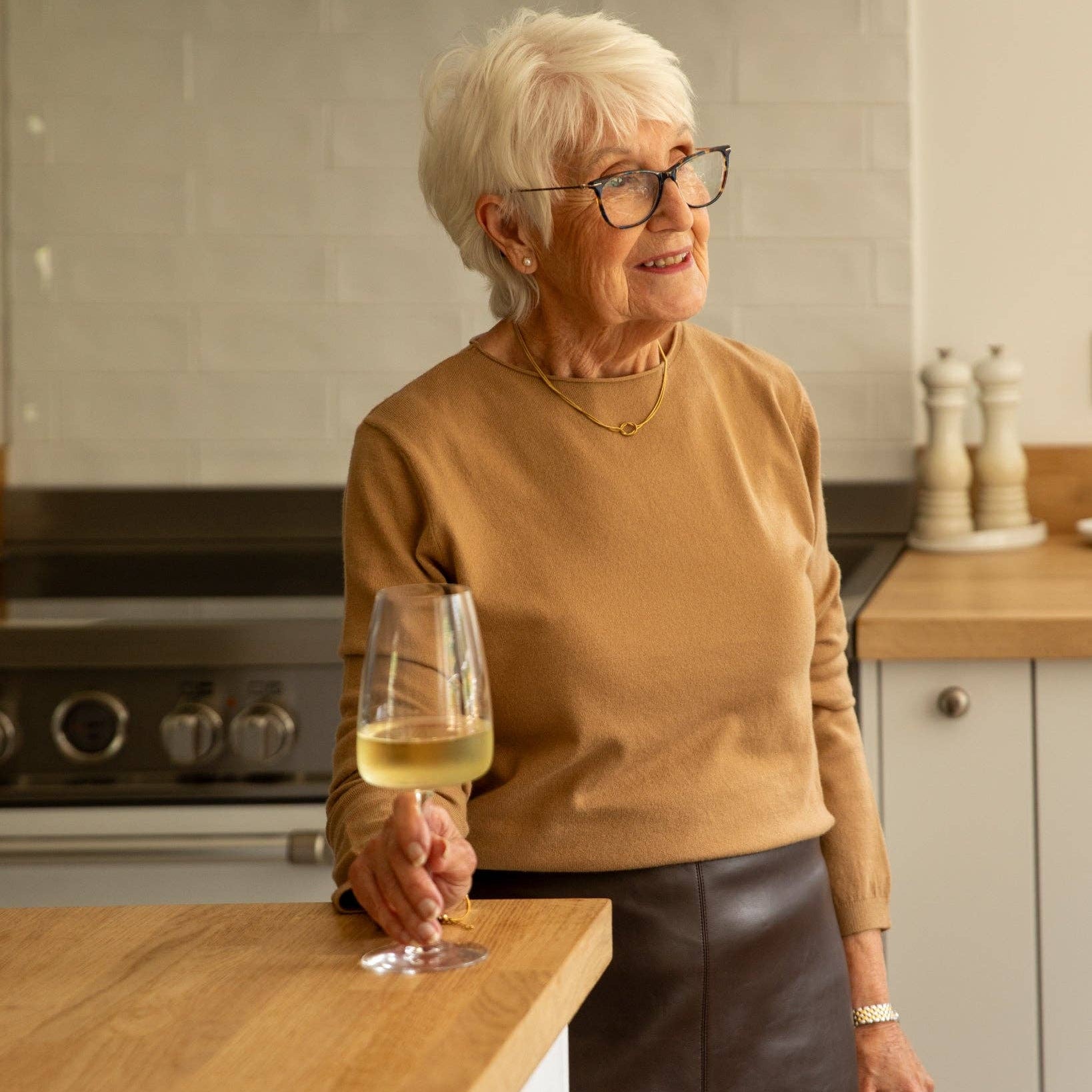 An older woman with short white hair and glasses stands in a kitchen, holding a glass of white wine. She is wearing a tan sweater and a dark skirt.
