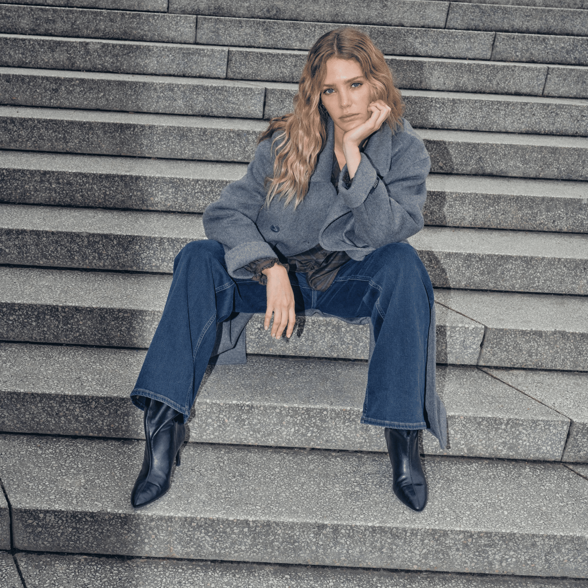 Woman sitting on stone steps wearing a gray coat and blue jeans.