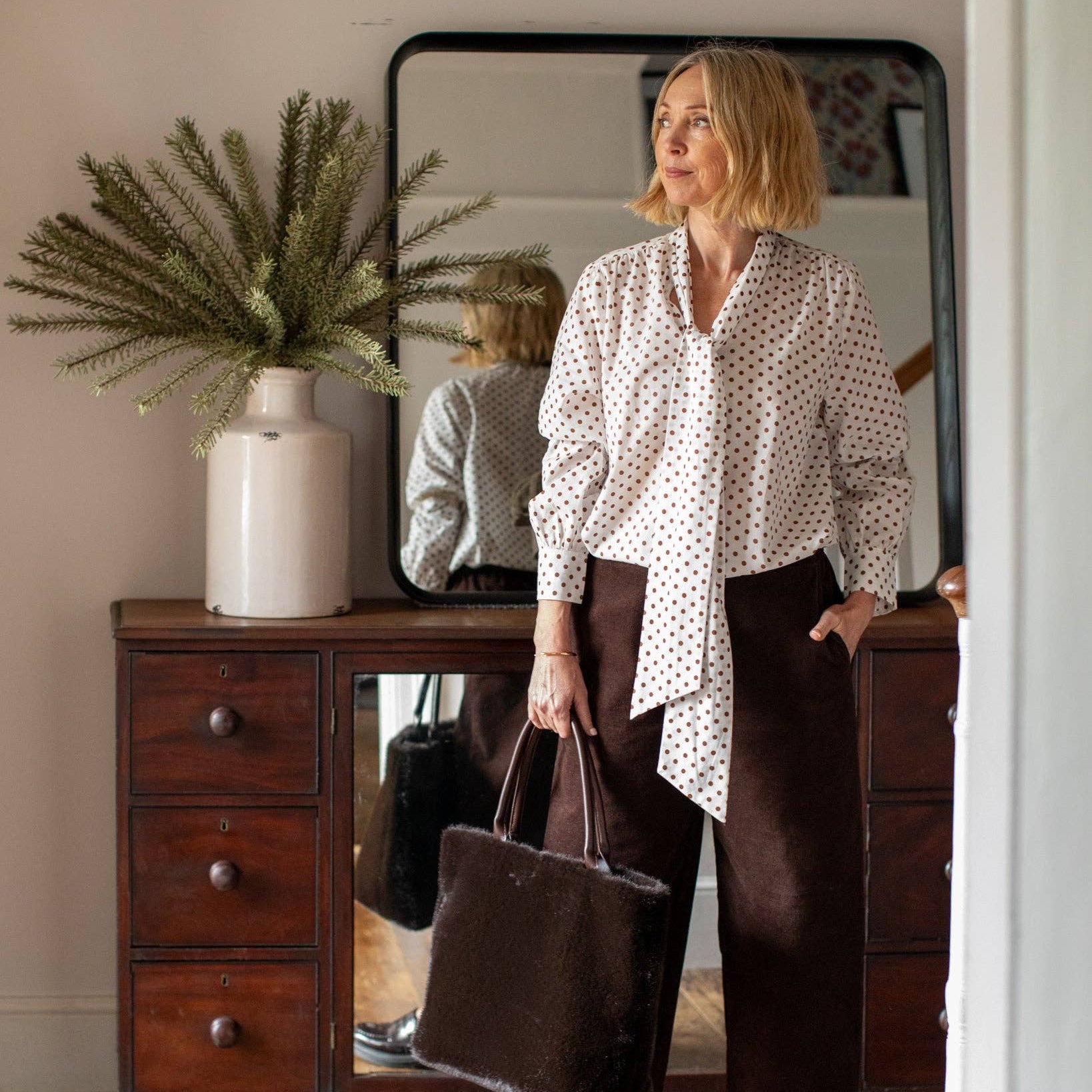 A woman stands in a room wearing a white button-up blouse with small black polka dots, dark brown wide-leg trousers, and black loafers, while holding a large dark brown tote bag. A wooden dresser, mirror, and potted plant are visible in the background.