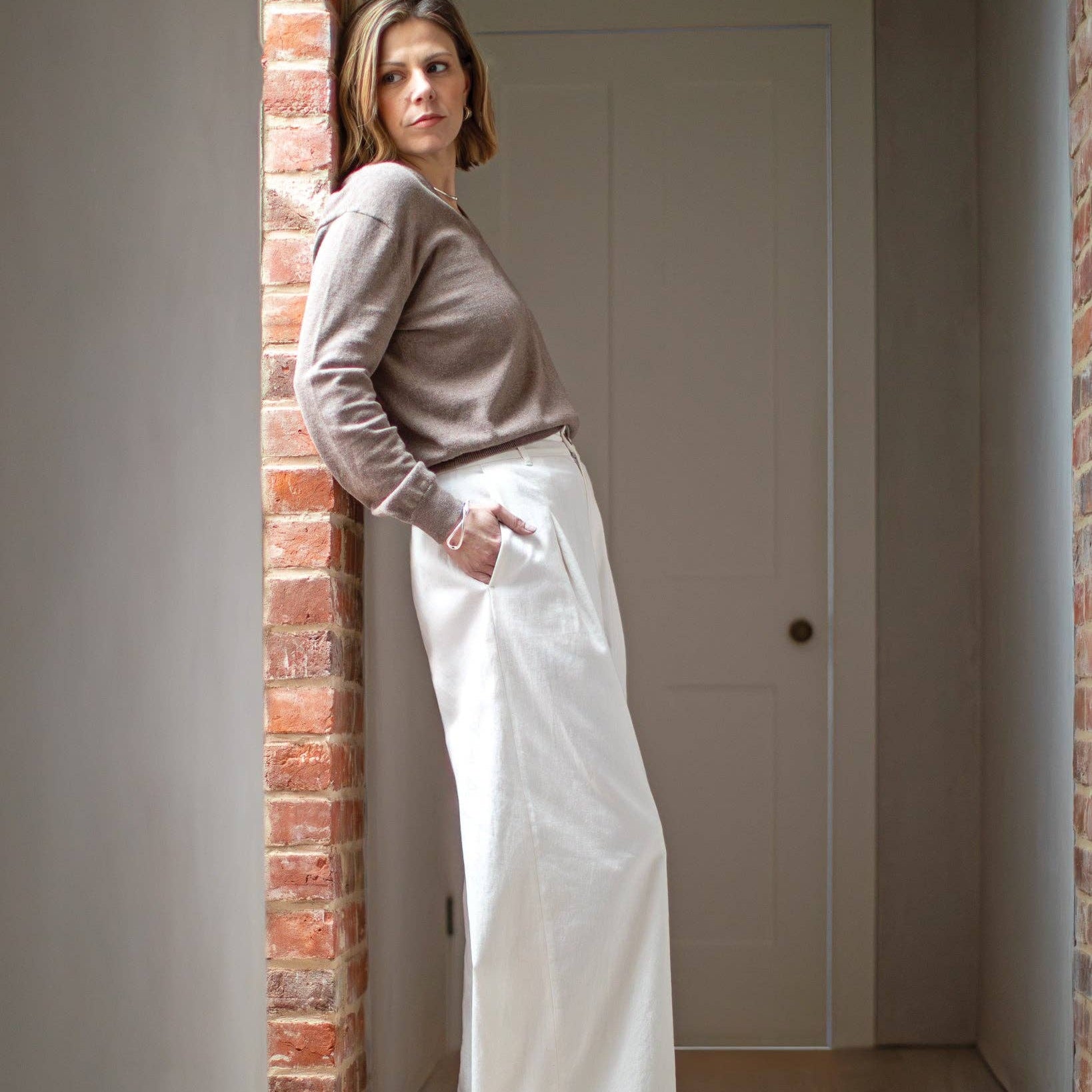 Woman in a beige sweater and white pants leaning against a brick wall.