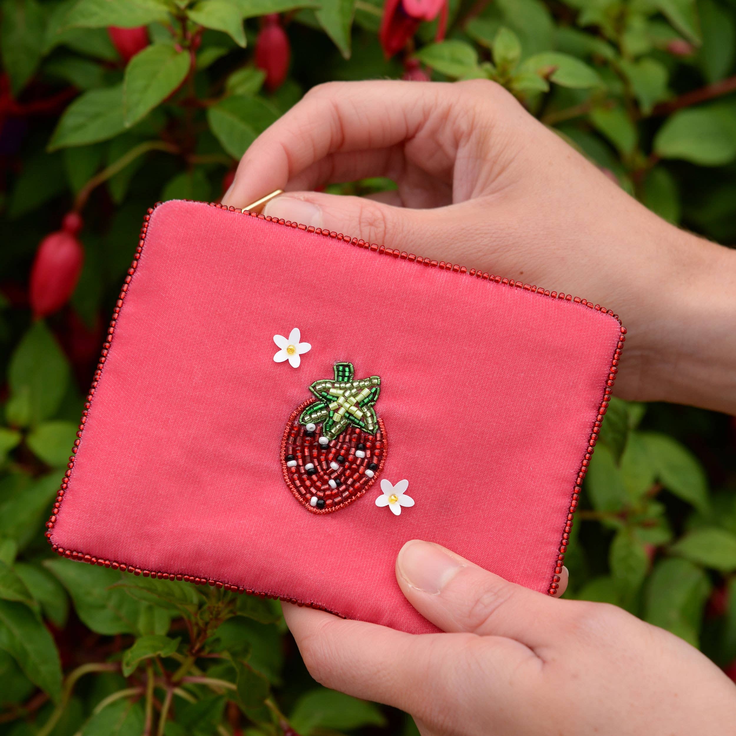 Hand holding a pink embroidered wallet by My Doris with flowers against a green leafy background