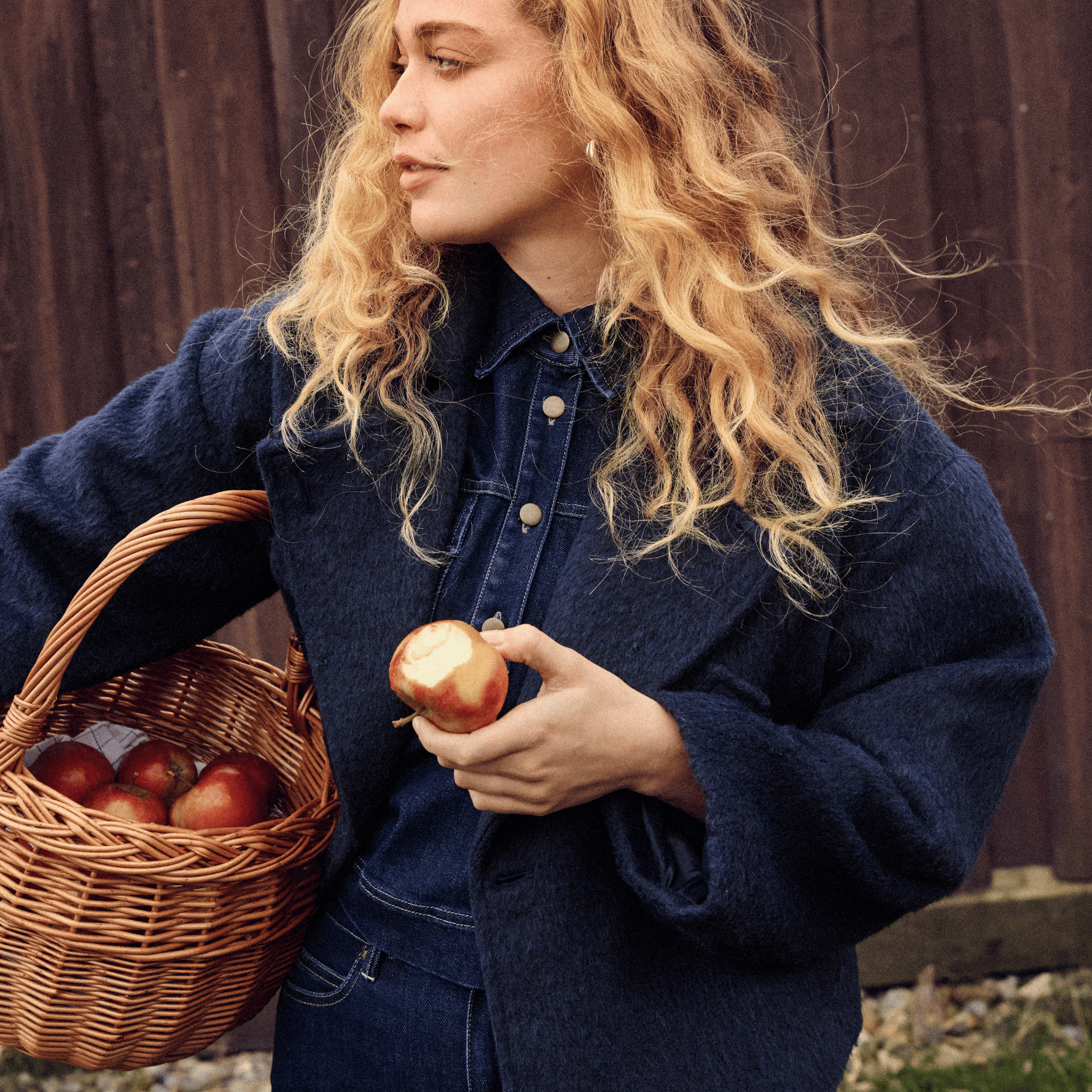 Woman in a dark blue wool coat holding a basket of apples and an apple in her hand against a wooden fence.