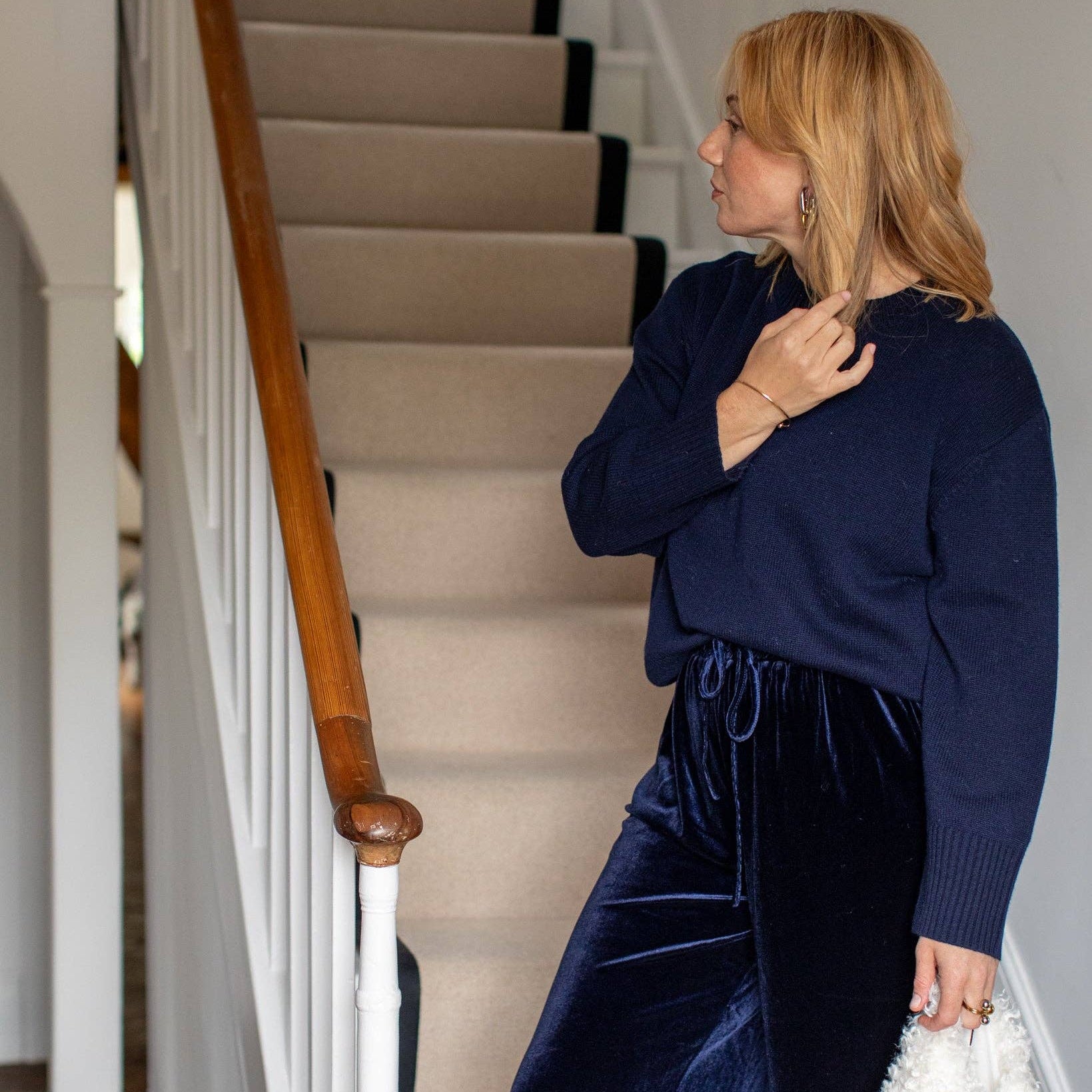A woman stands on an indoor staircase wearing a navy blue knit sweater and matching navy velvet drawstring pants, holding a white textured handbag.