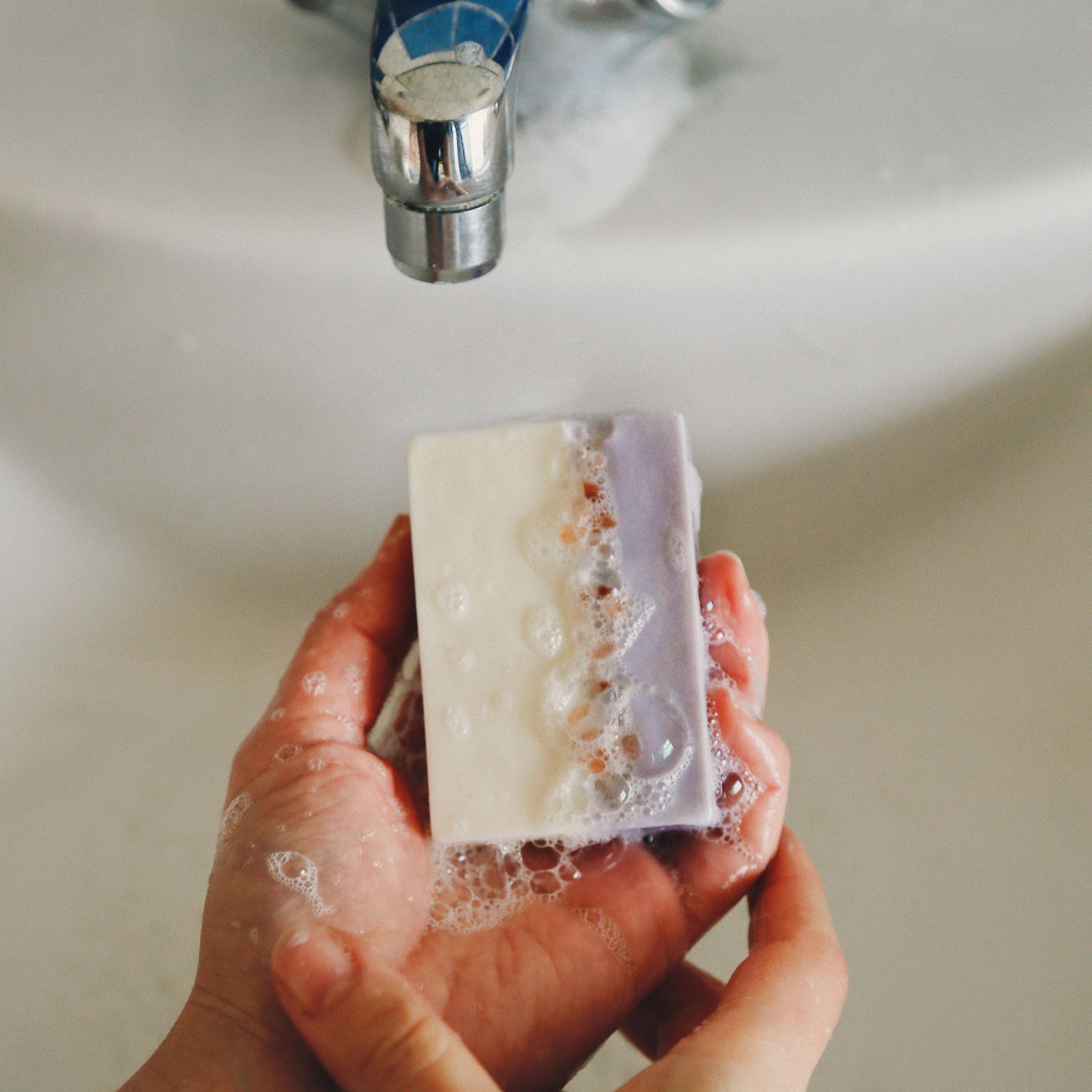 Hand holding a bar of soap under running water from a faucet.