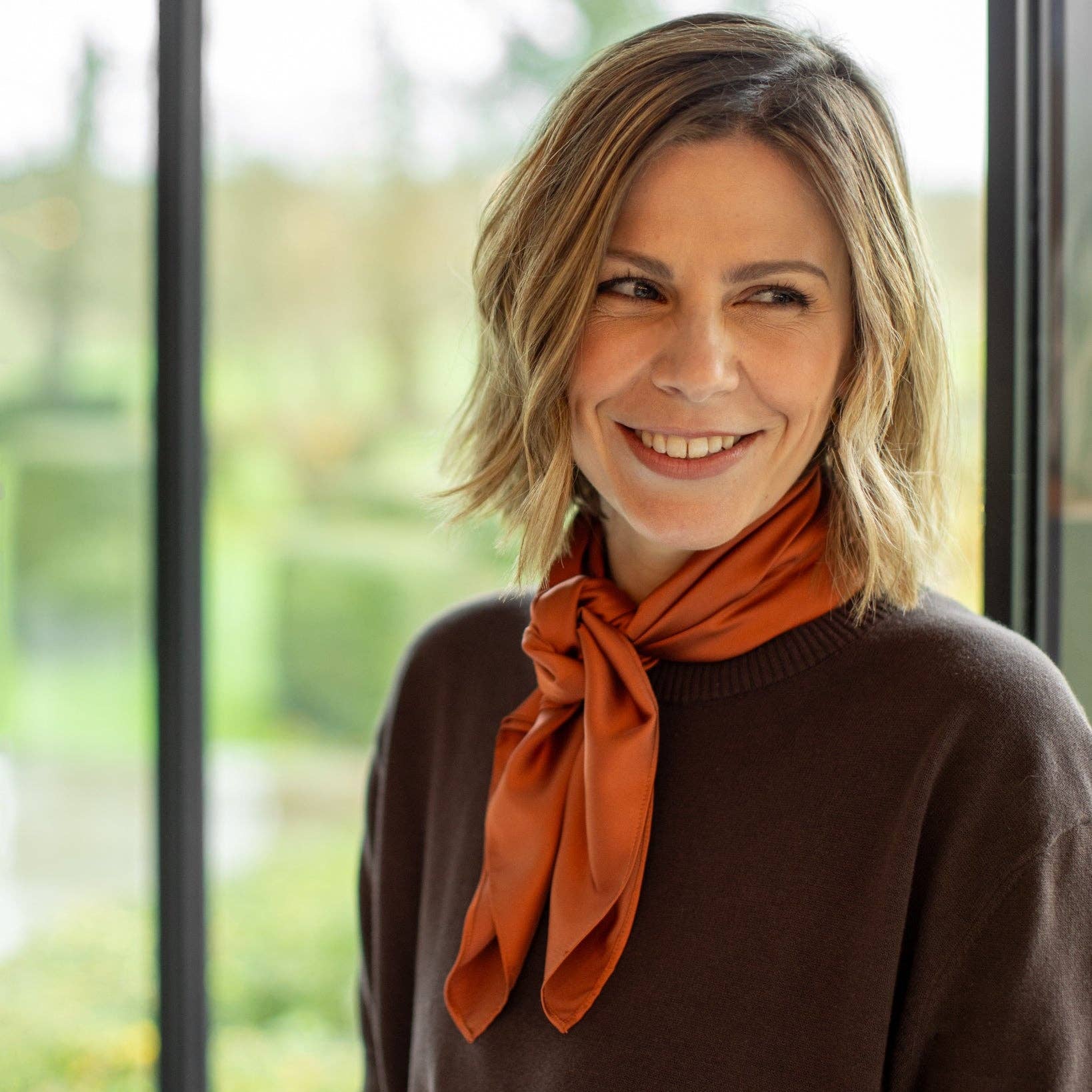 Woman wearing a brown sweater and orange scarf standing indoors with a blurred green outdoor background