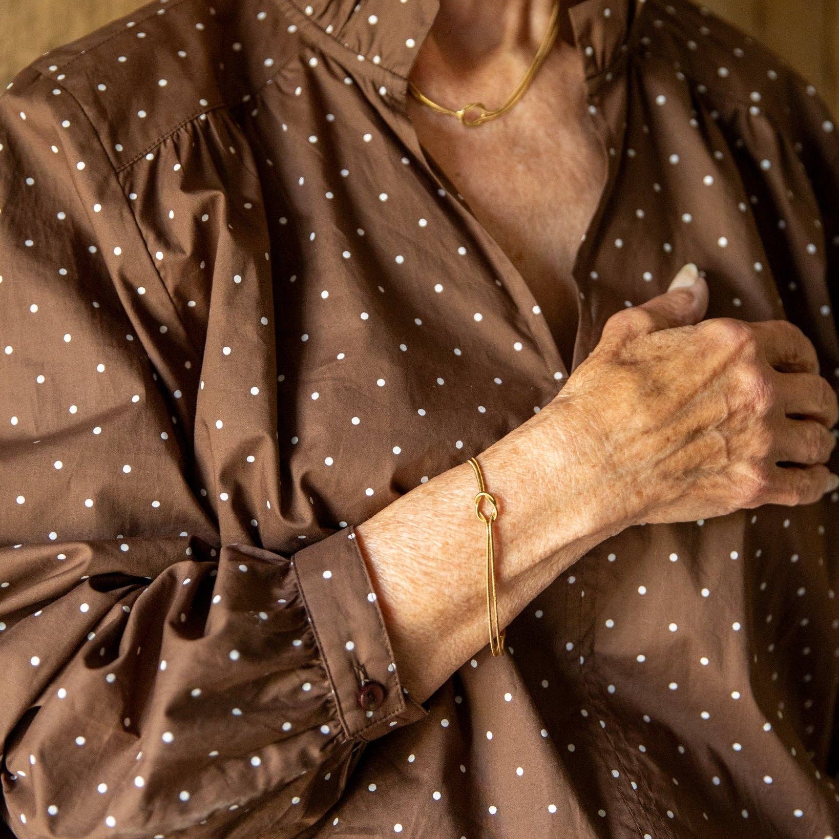 A close-up of a person wearing a brown long-sleeve button-up shirt with a white polka dot pattern, paired with dark blue jeans. The person is also wearing a delicate gold chain necklace and a gold chain bracelet.