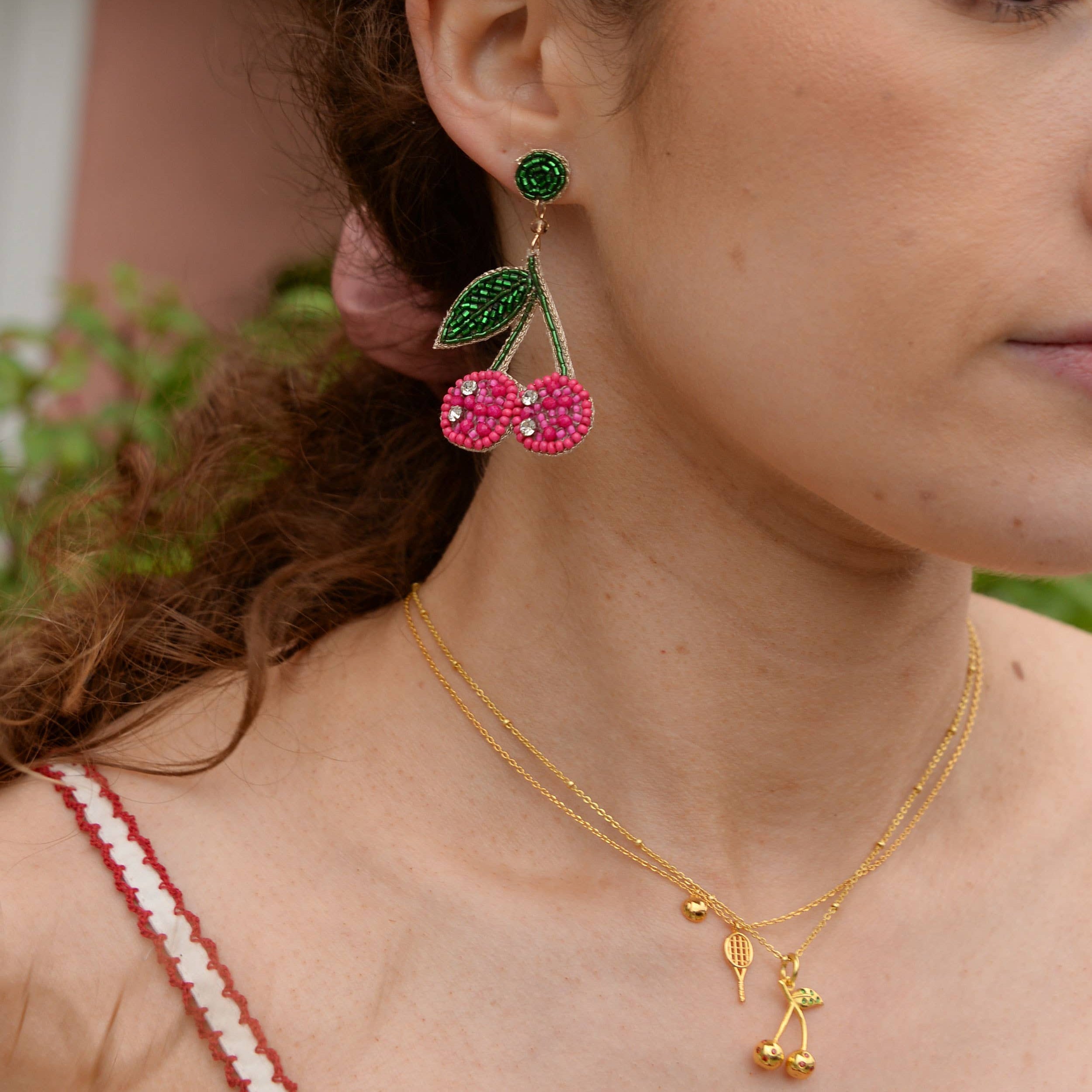 Close-up of a woman wearing colorful earrings and a gold necklace with a cherry pendant.