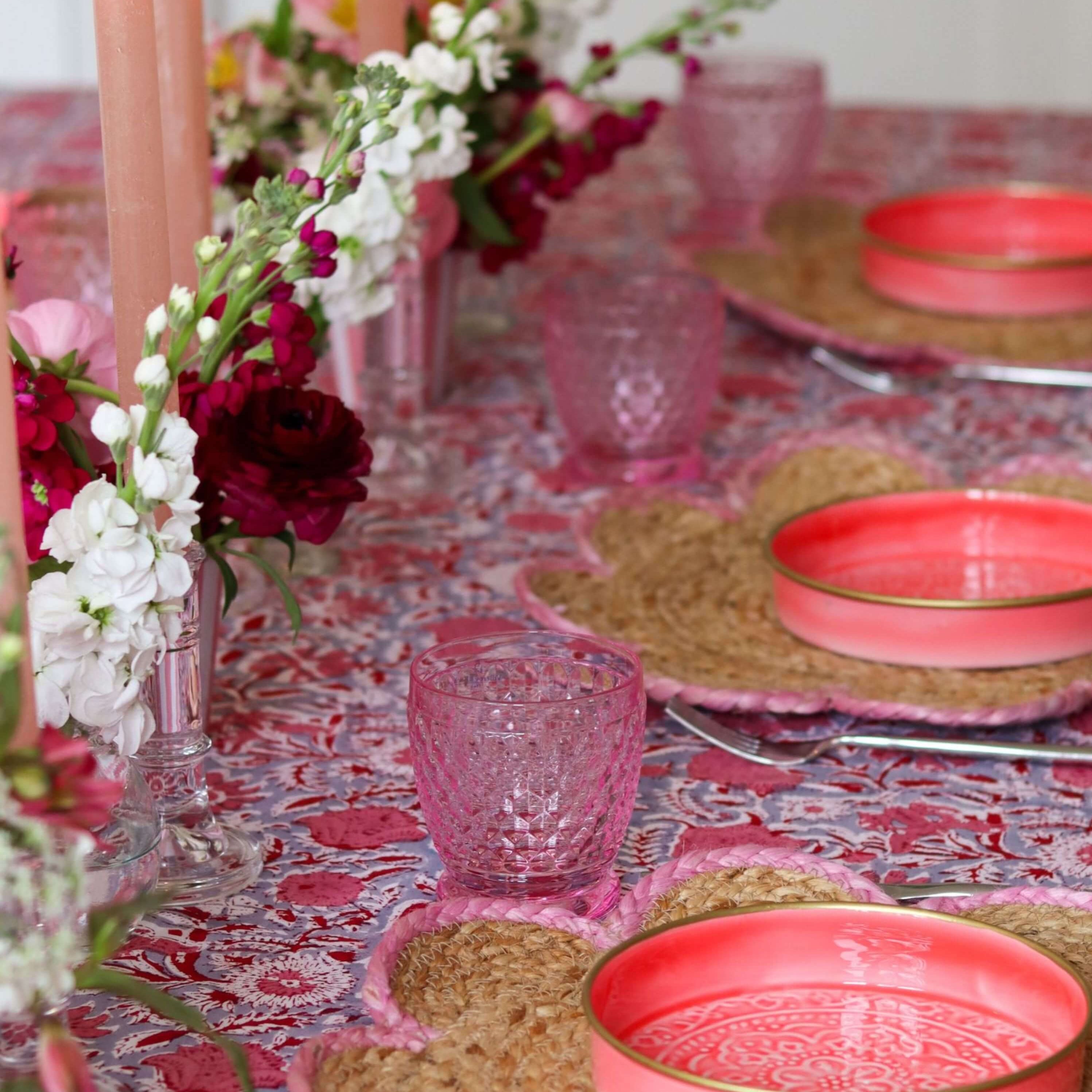 Decorative table setting with pink plates, glasses, and flowers on a patterned tablecloth.