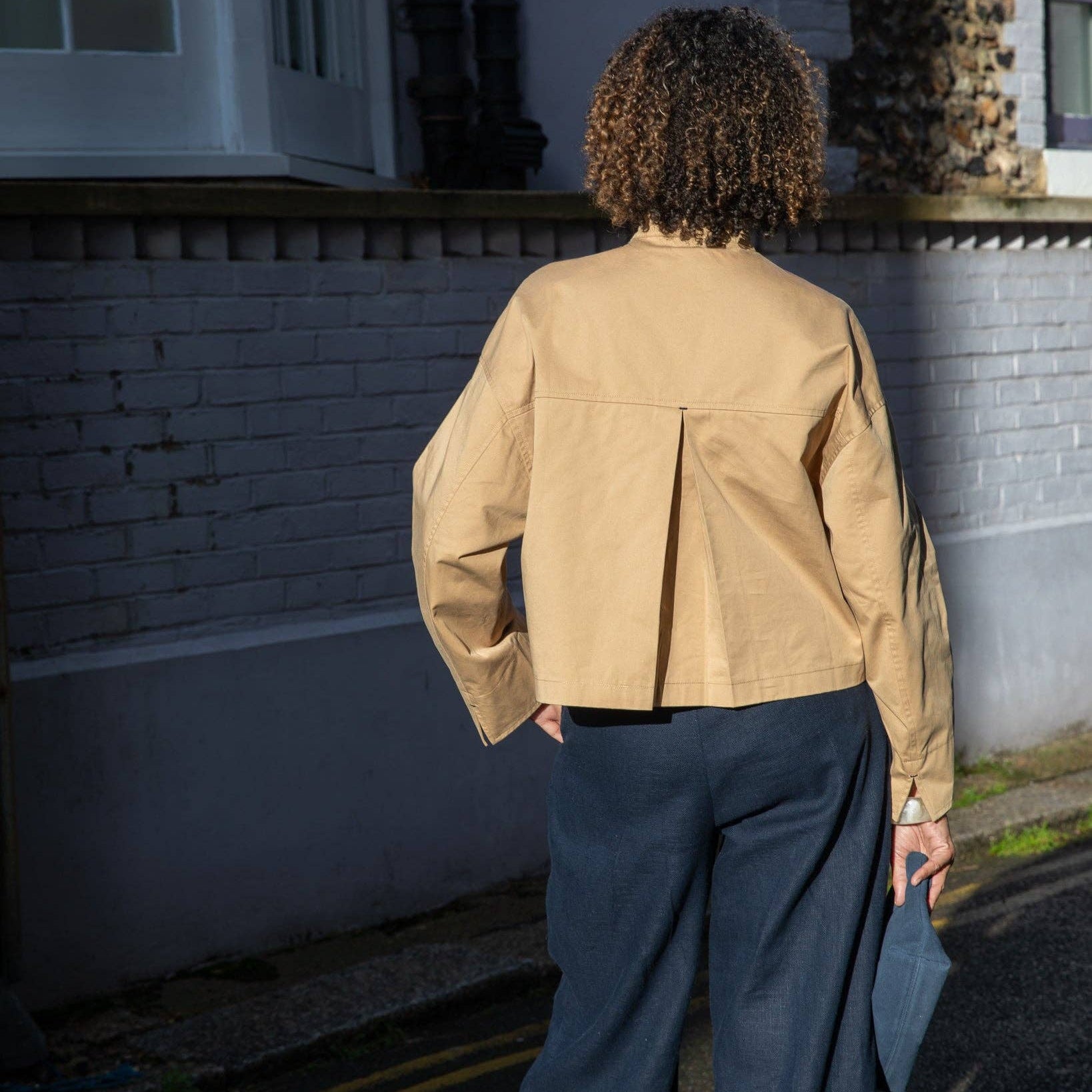 Person wearing a beige jacket and dark pants standing on a street.