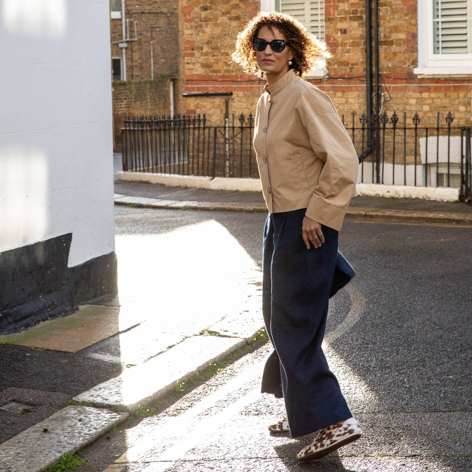 Woman walking on a street wearing sunglasses and a beige jacket.