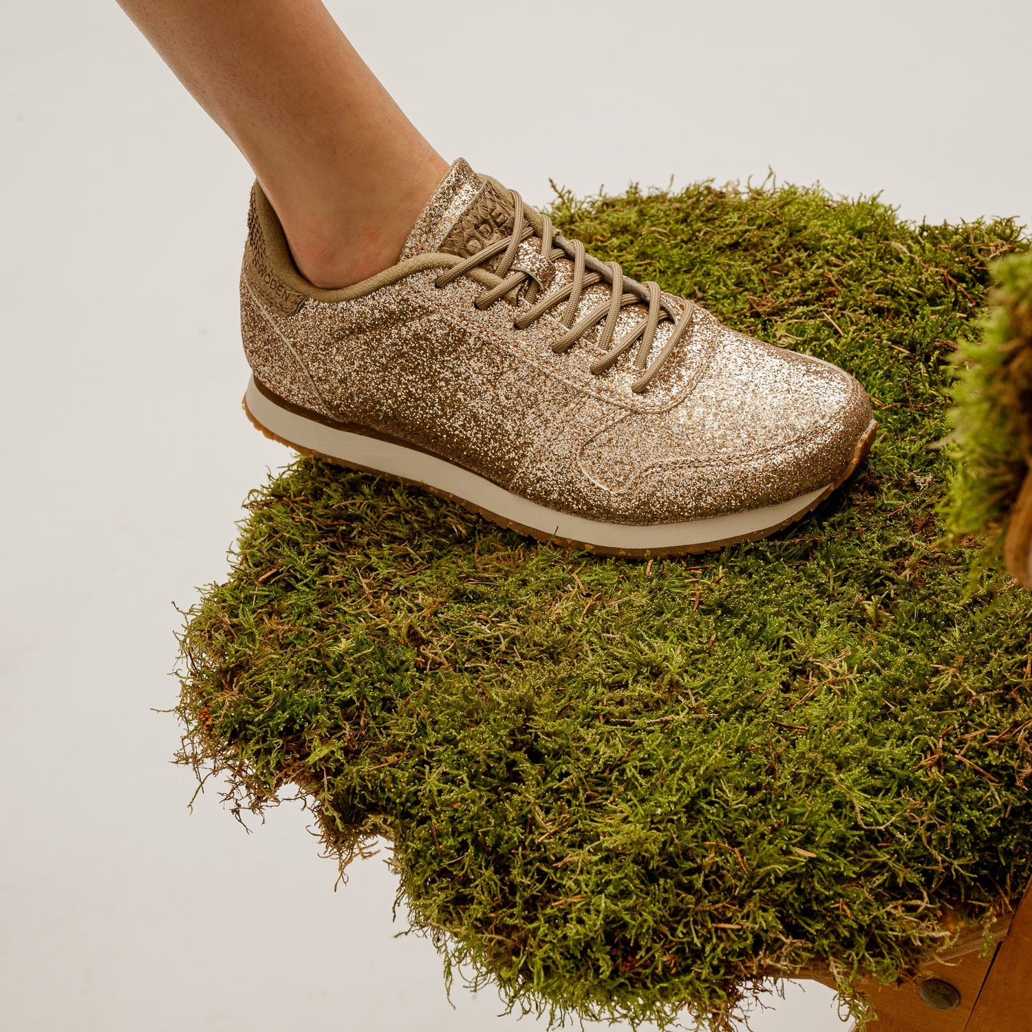 A gold glitter-covered sneaker with a rubber heel cap and leather details, placed on a bed of moss.