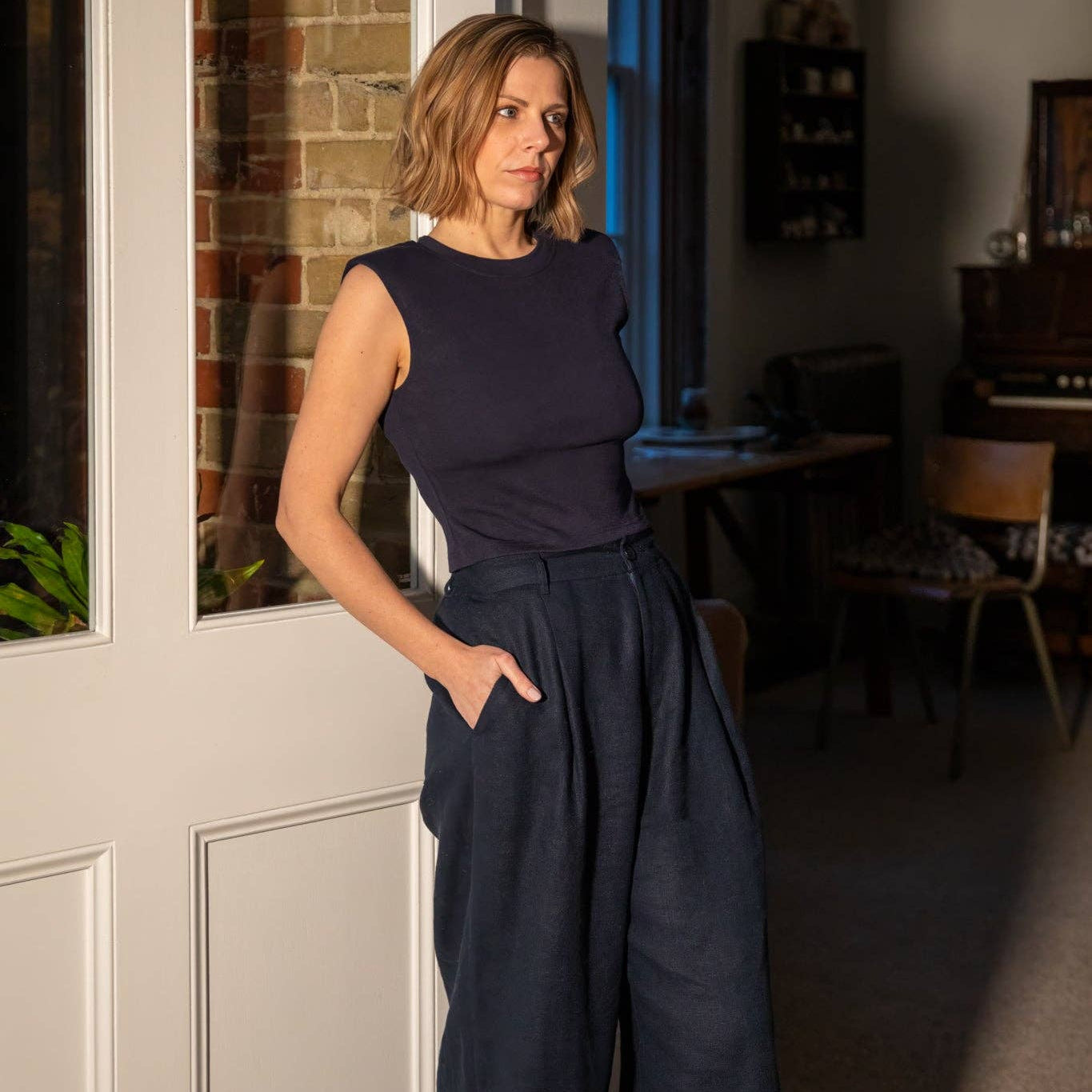 Woman wearing a navy sleeveless top and wide-leg pants standing in a room with a door and chair.