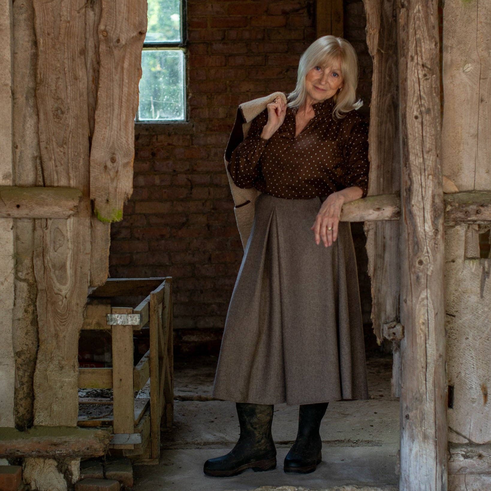 An older woman with blonde hair stands in a rustic wooden doorway of a brick building, wearing a dark brown patterned blouse, a long taupe skirt, and black boots.