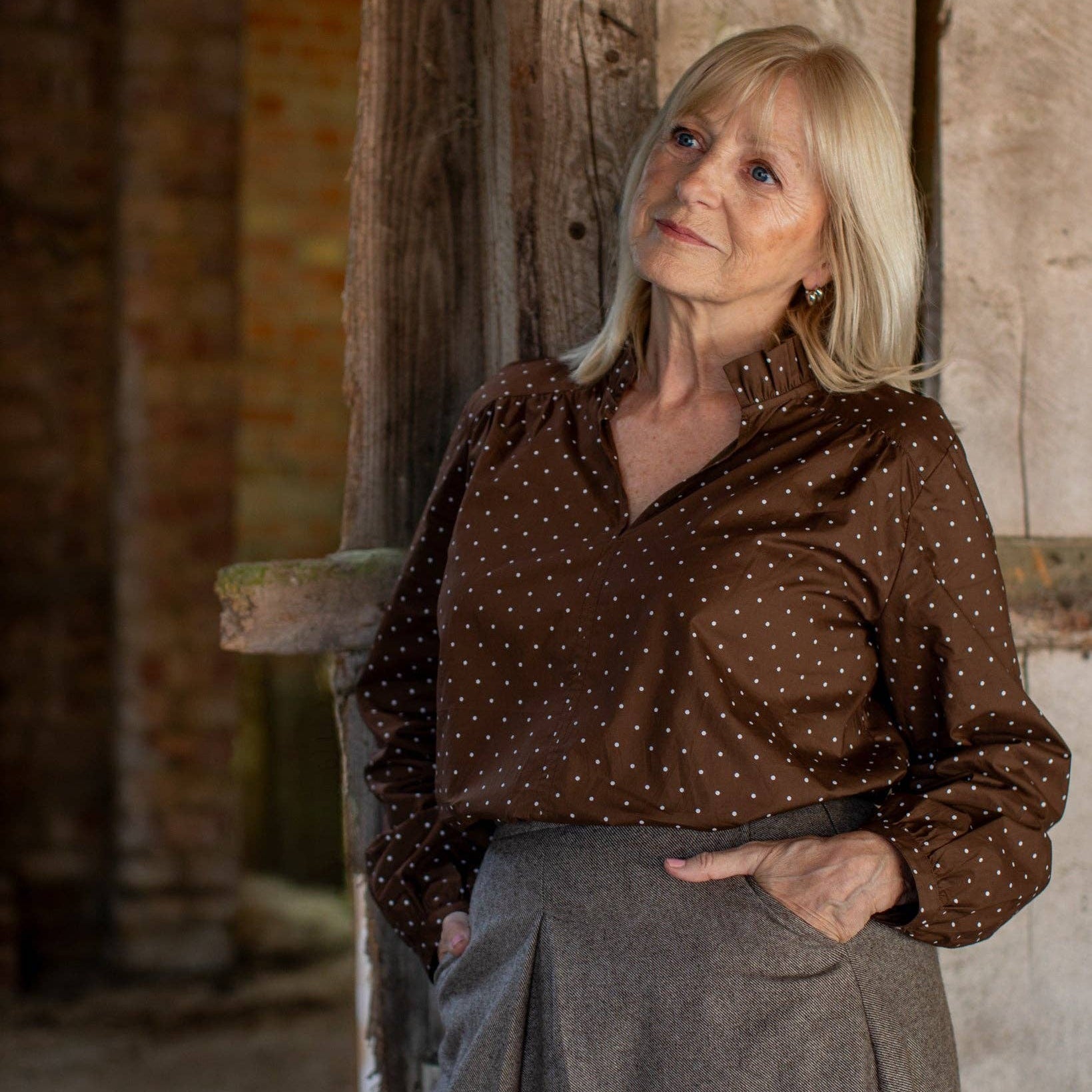 A woman with blonde hair stands in a rustic indoor setting, wearing a brown long-sleeve blouse with small white polka dots and a gray skirt.