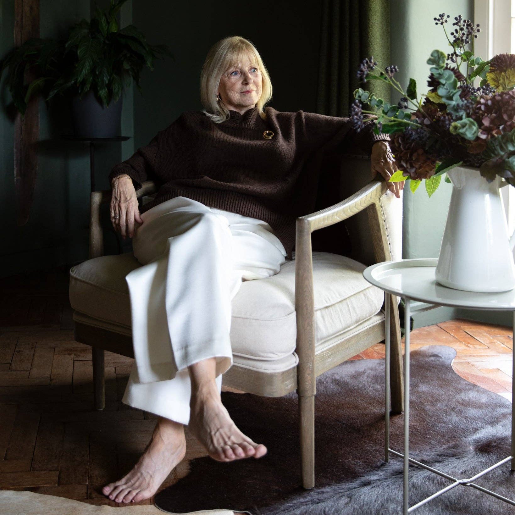 A woman sits barefoot in a cream upholstered armchair in a dark, stylish room. She is wearing a dark brown mock-neck knit sweater and white wide-leg trousers.