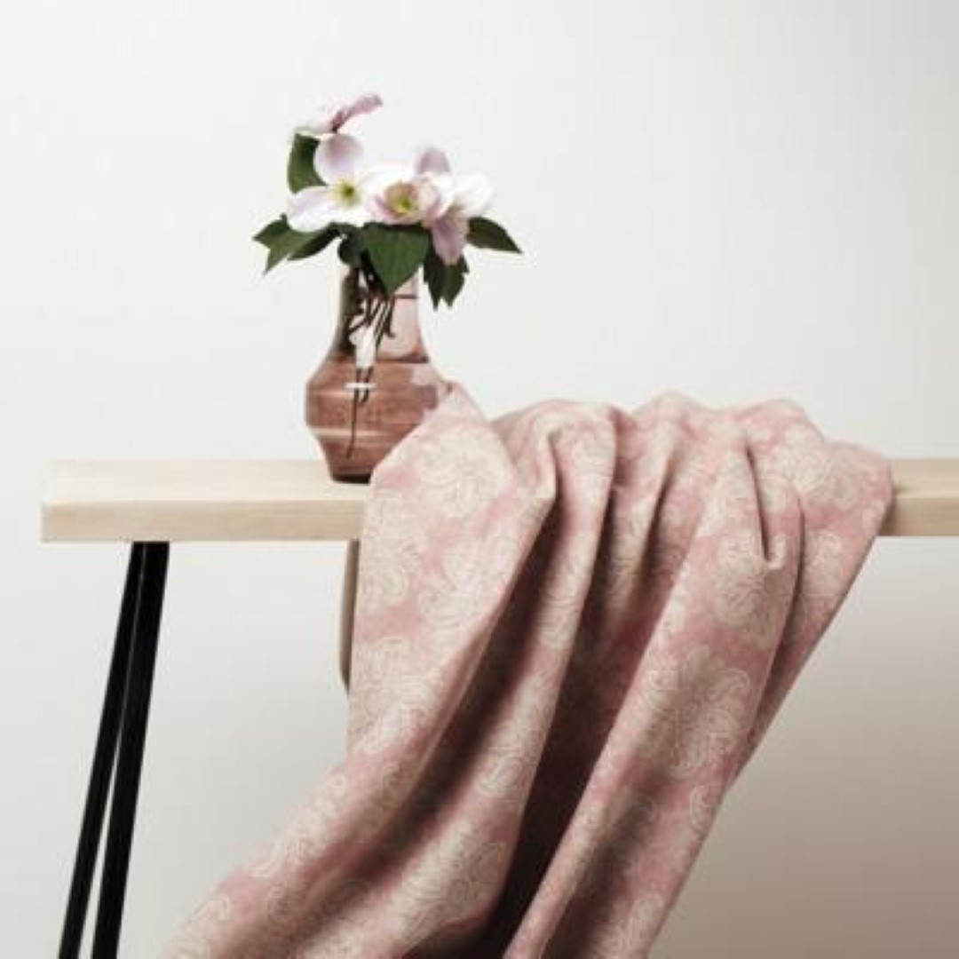 A fabric with a floral pattern draped over a table, in shades of pink, with a vase of flowers in the background.