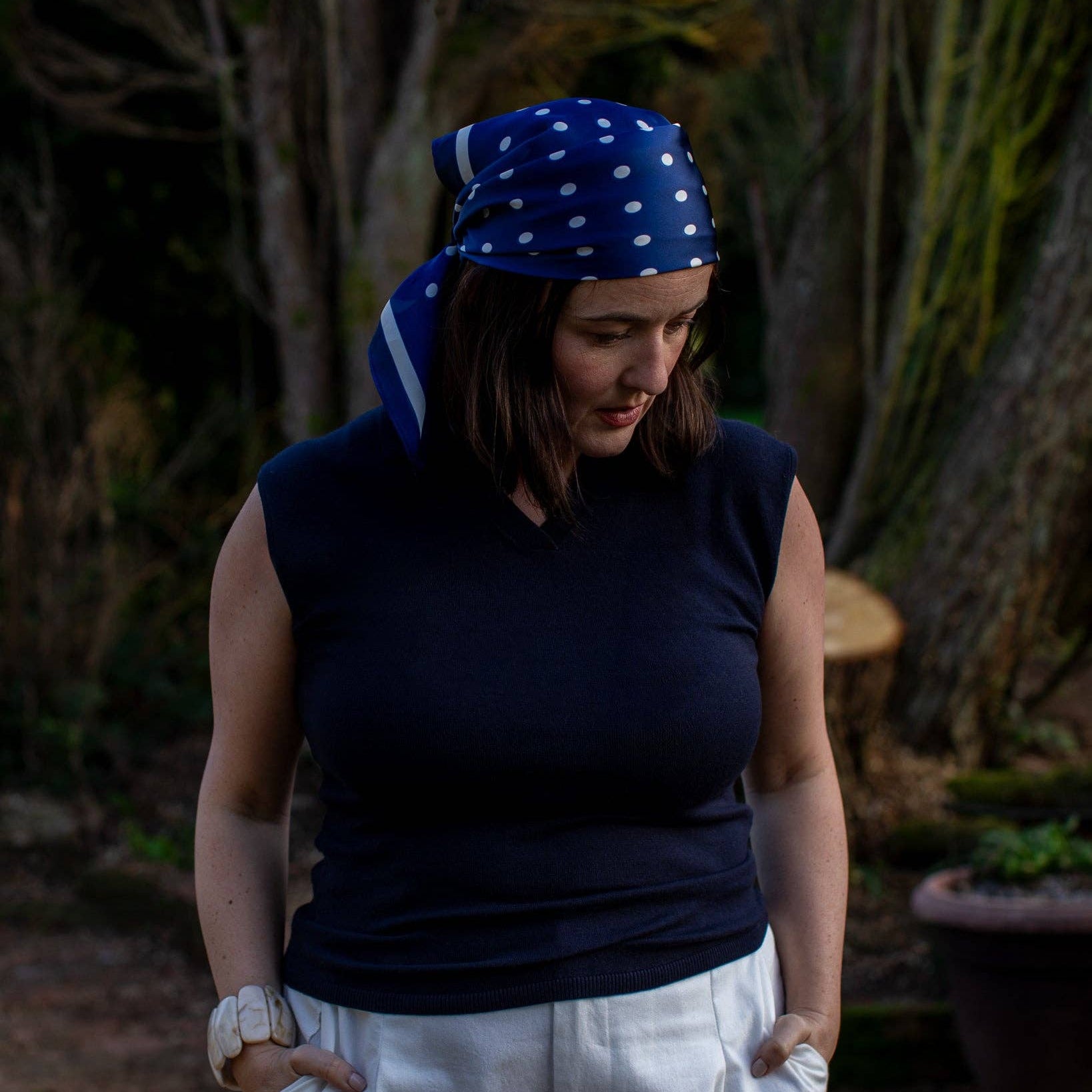 Woman wearing a navy sleeveless top and white pants with a blue polka dot headscarf outdoors.