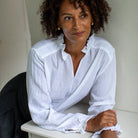 A woman with curly dark hair wearing a white long-sleeve button-up shirt with ruffled trim, leaning on a white table against a light wall.