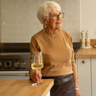 An older woman with short white hair and glasses stands in a kitchen, holding a glass of white wine. She is wearing a tan sweater and a dark skirt.
