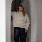 A woman wearing a cream ribbed turtleneck sweater and dark wide-leg trousers stands in a white arched hallway.