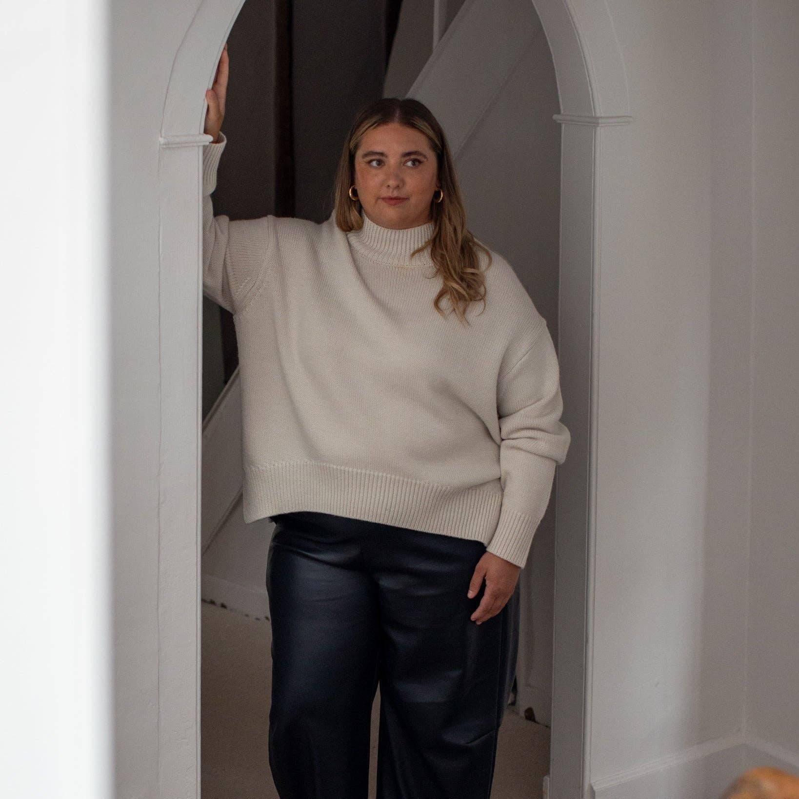 A woman wearing a cream ribbed turtleneck sweater and dark wide-leg trousers stands in a white arched hallway.