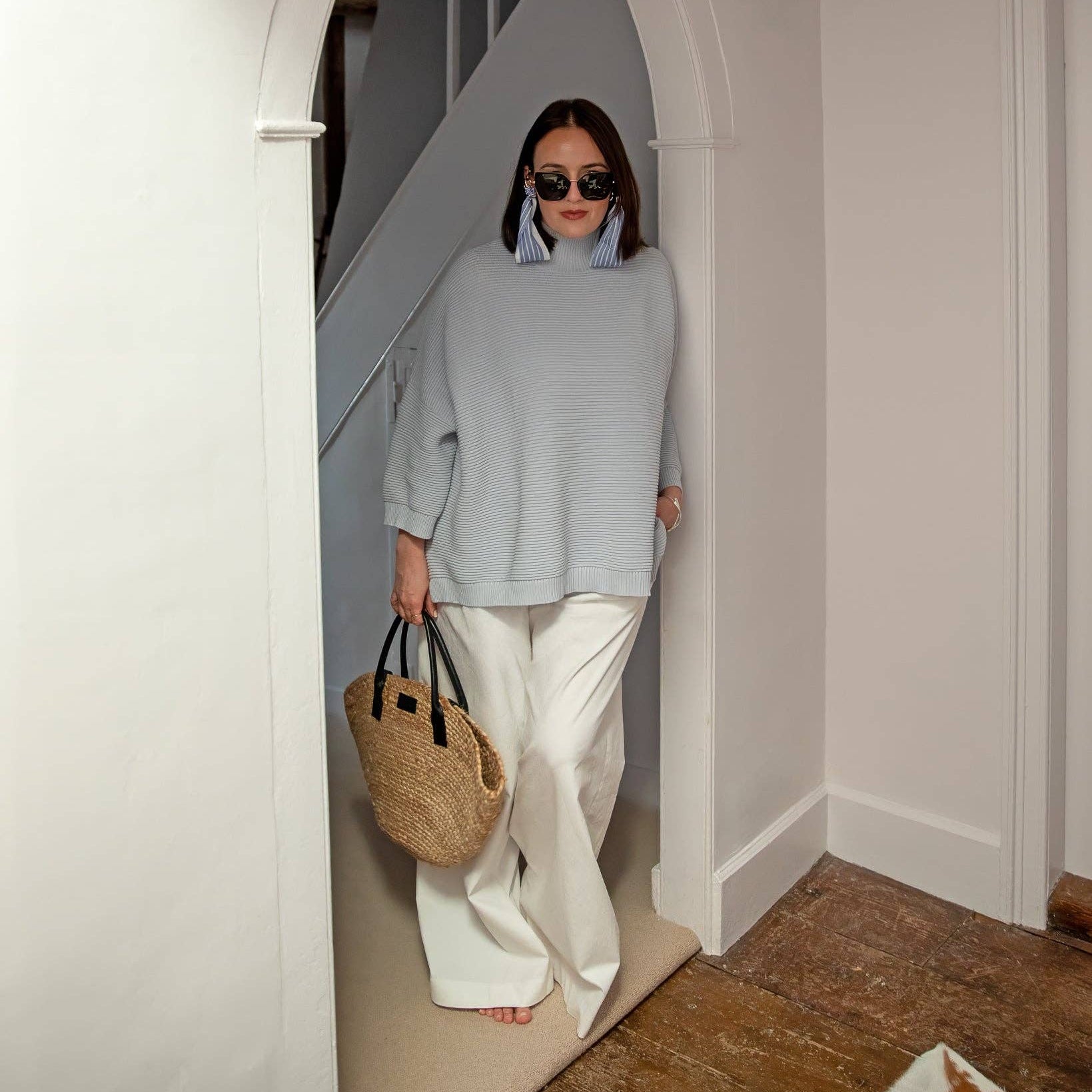 Person wearing a light blue sweater and white pants, holding a straw bag, standing in a home interior.
