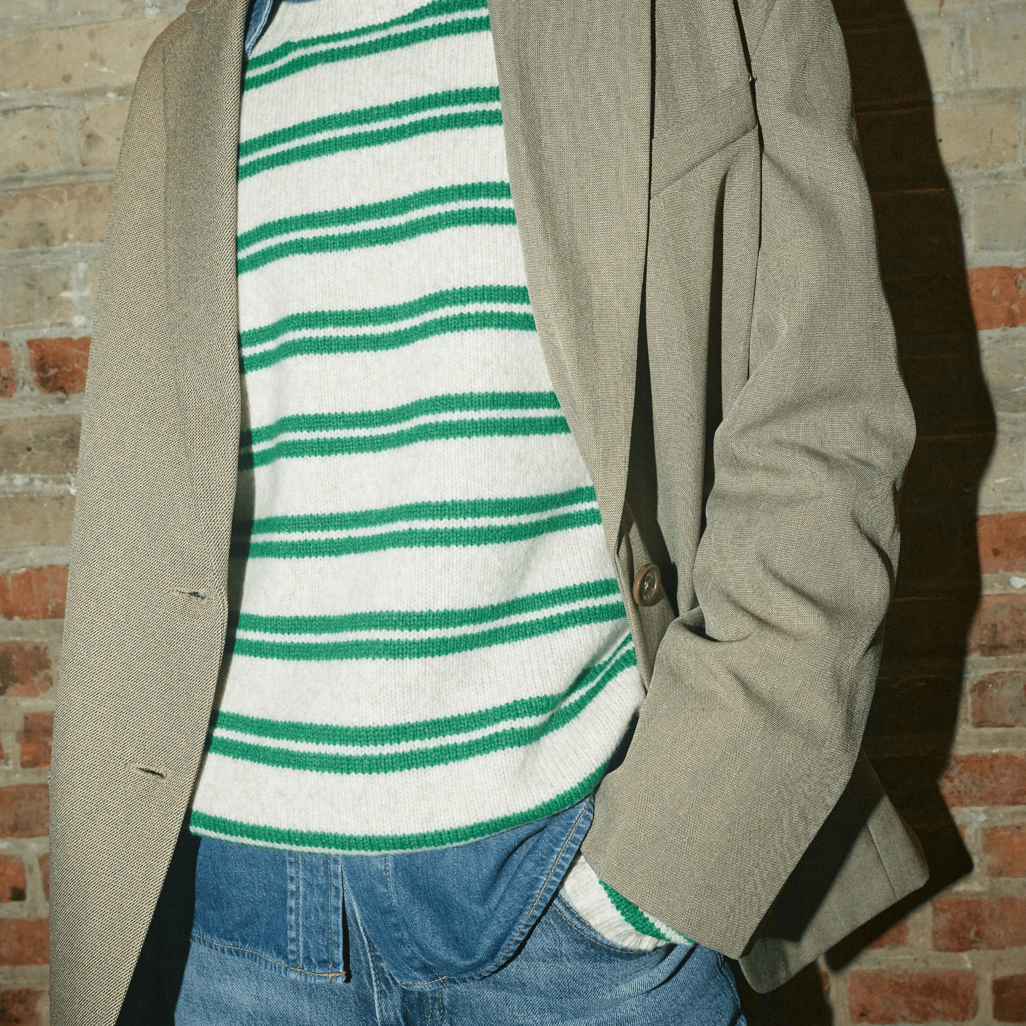 Person wearing a beige coat over a green and white striped shirt with blue jeans, standing against a brick wall.