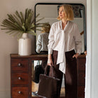 A woman stands in a room wearing a white button-up blouse with small black polka dots, dark brown wide-leg trousers, and black loafers, while holding a large dark brown tote bag. A wooden dresser, mirror, and potted plant are visible in the background.