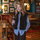 A woman standing in a cozy, dimly lit pub or restaurant, wearing a brown faux-fur vest over a light blue button-down shirt and dark trousers, holding a glass of red wine.