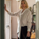 A woman with blonde hair wearing a white button-down shirt with a black polka-dot pattern and dark brown trousers, standing in a doorway and holding a black handbag.
