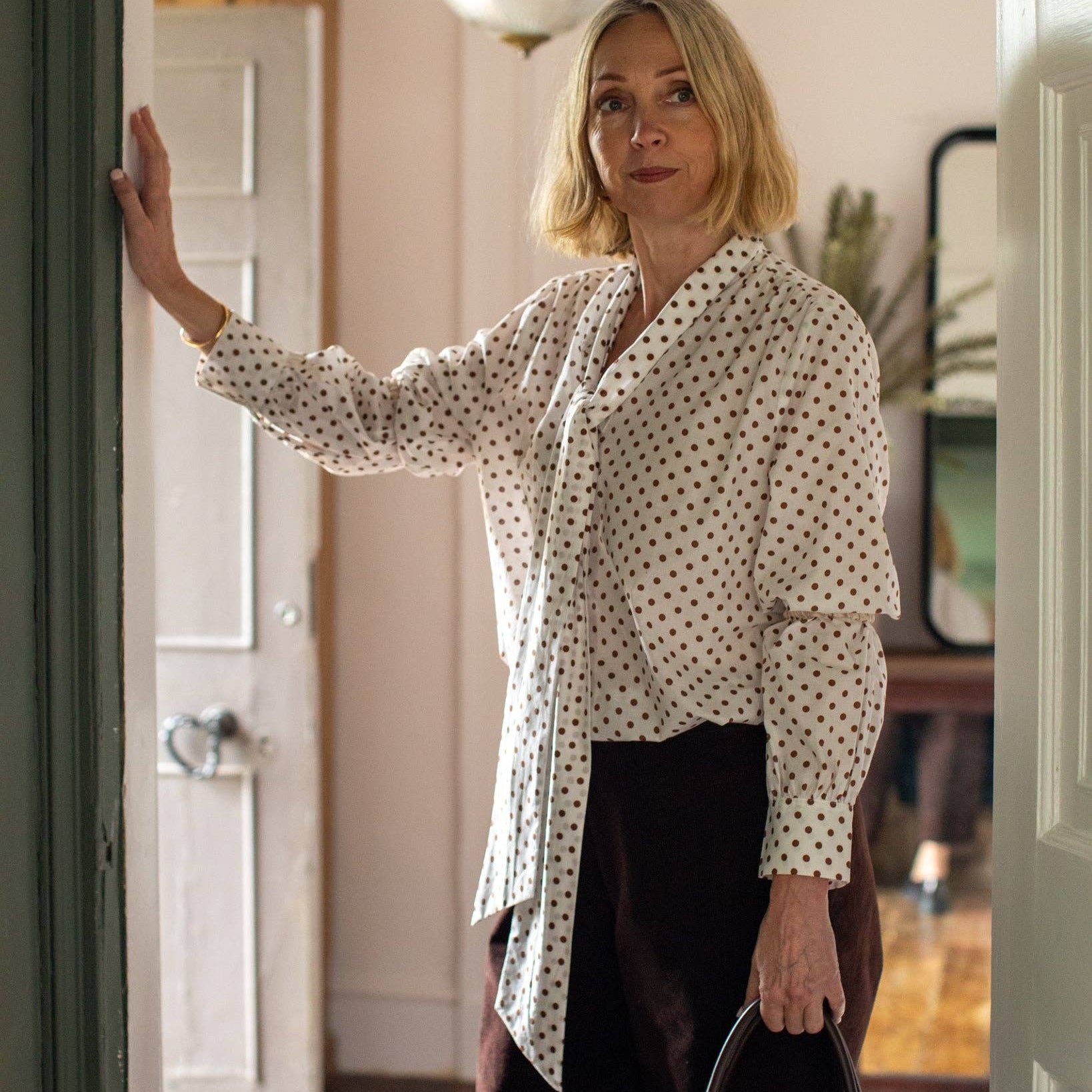 A woman with blonde hair wearing a white button-down shirt with a black polka-dot pattern and dark brown trousers, standing in a doorway and holding a black handbag.