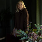 A woman with blonde hair wearing a dark brown turtleneck sweater stands in a dimly lit room, with a floral arrangement in the foreground and a striped armchair behind her.
