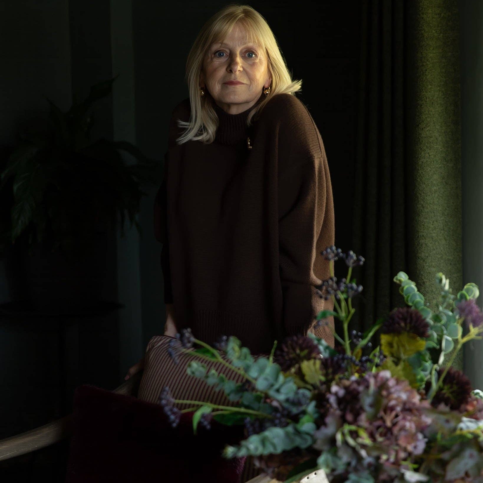 A woman with blonde hair wearing a dark brown turtleneck sweater stands in a dimly lit room, with a floral arrangement in the foreground and a striped armchair behind her.