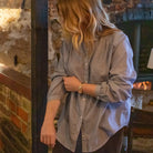 A woman with blonde hair wearing a light blue cotton button-up shirt and dark brown trousers, standing in a rustic room with a brick wall and a fireplace in the background.