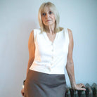Woman wearing a white sleeveless top and gray skirt standing next to a radiator against a plain wall.