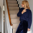A woman stands on an indoor staircase wearing a navy blue knit sweater and matching navy velvet drawstring pants, holding a white textured handbag.