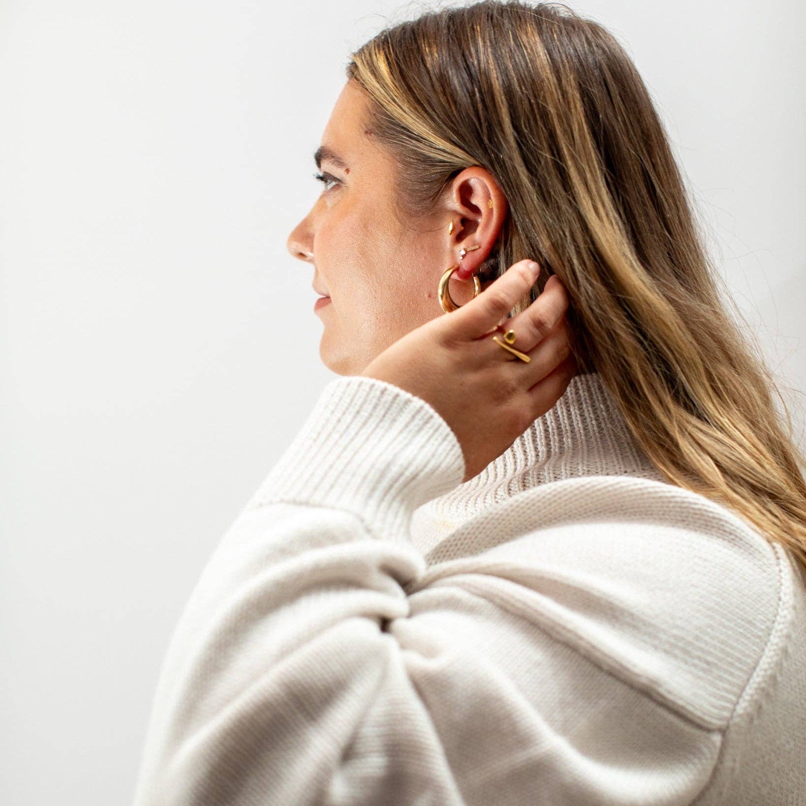 A woman in profile wearing a cream ribbed knit sweater and gold hoop earrings, shown against a plain light gray background.