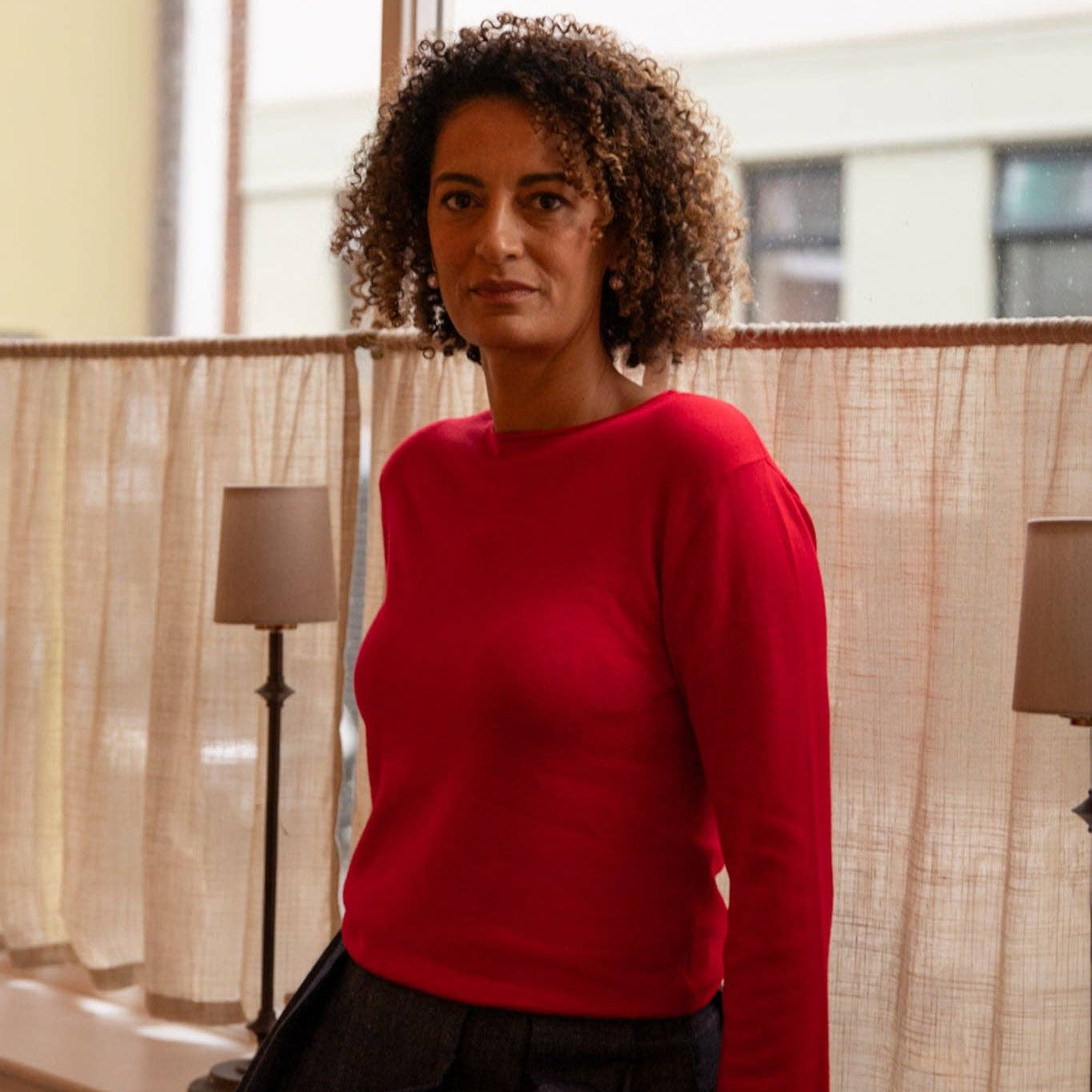 Woman wearing a red sweater and black pants standing in a room with curtains and lamps.