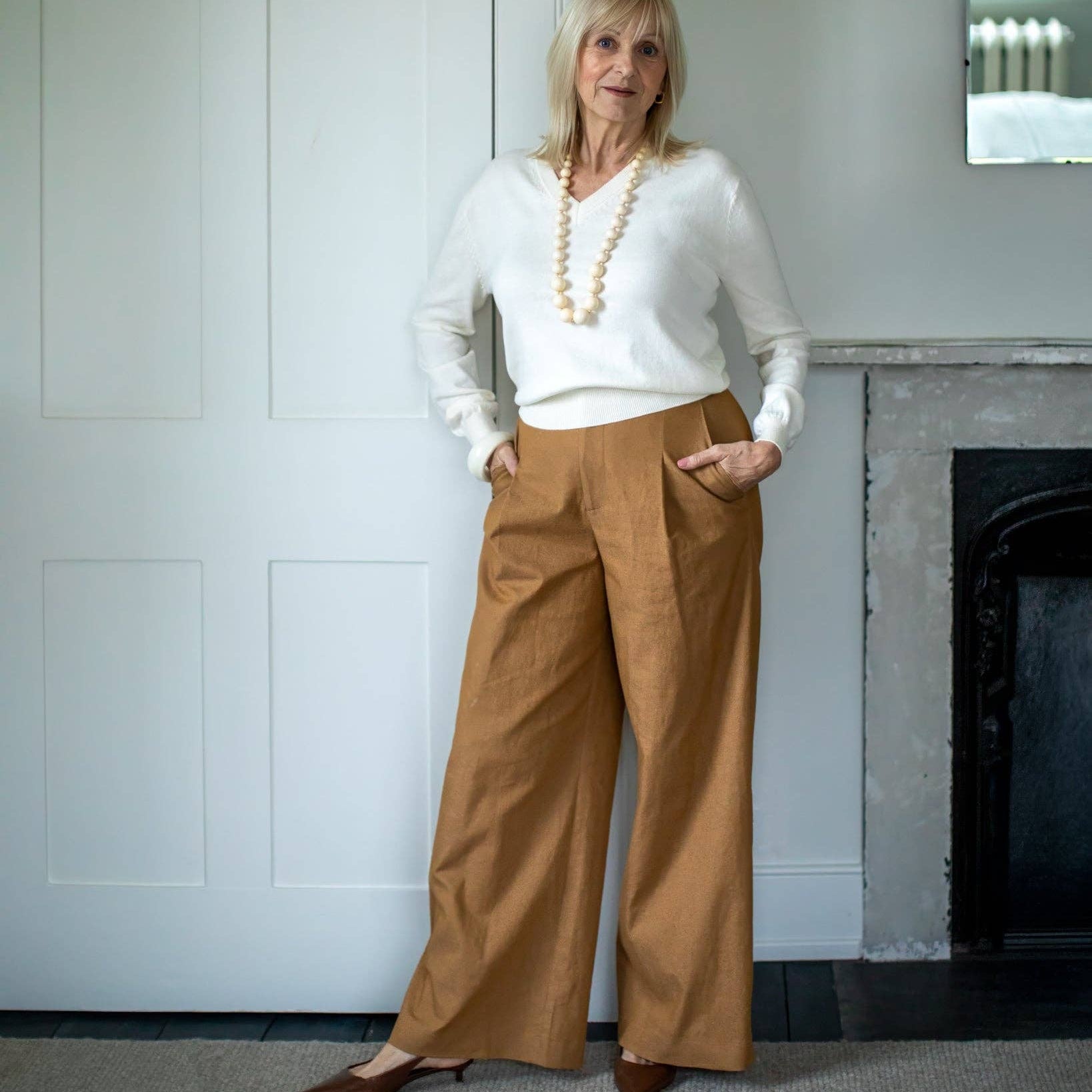 Woman wearing a white sweater and brown pants standing in a room with a fireplace.