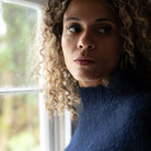 A close-up portrait of a woman with curly blonde hair wearing a fuzzy navy blue mock-neck sweater, standing near a window.