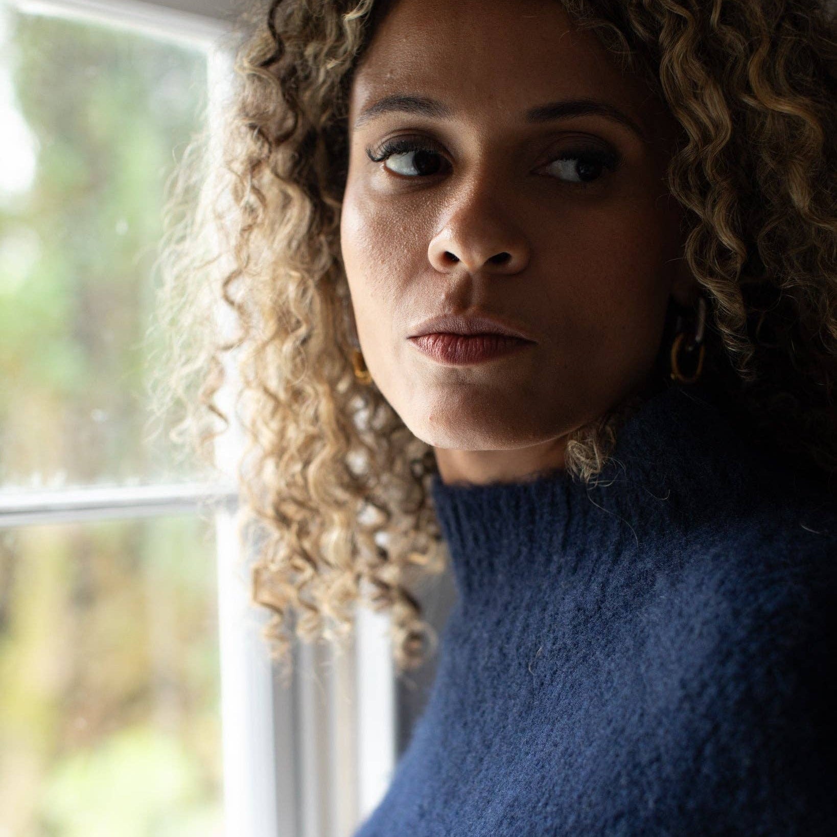 A close-up portrait of a woman with curly blonde hair wearing a fuzzy navy blue mock-neck sweater, standing near a window.