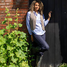A woman with curly hair stands outdoors against a dark wooden door and brick wall, wearing a light blue button-up shirt, taupe vest, dark pants, and green rubber boots. Large green plants are visible in the foreground.