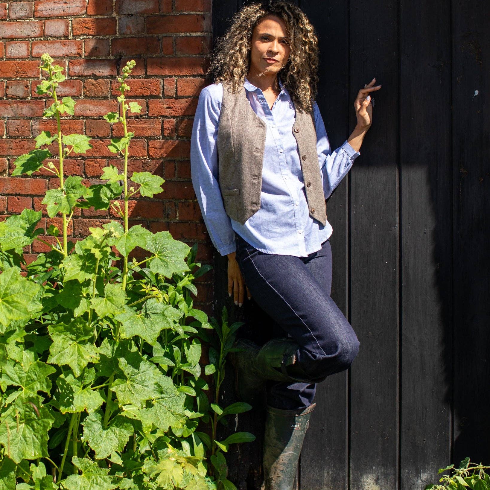 A woman with curly hair stands outdoors against a dark wooden door and brick wall, wearing a light blue button-up shirt, taupe vest, dark pants, and green rubber boots. Large green plants are visible in the foreground.