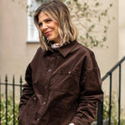 A woman wearing a dark brown corduroy button-up jacket and matching brown trousers, with cream spotty neck scarf standing outdoors against a beige wall with a black metal railing.