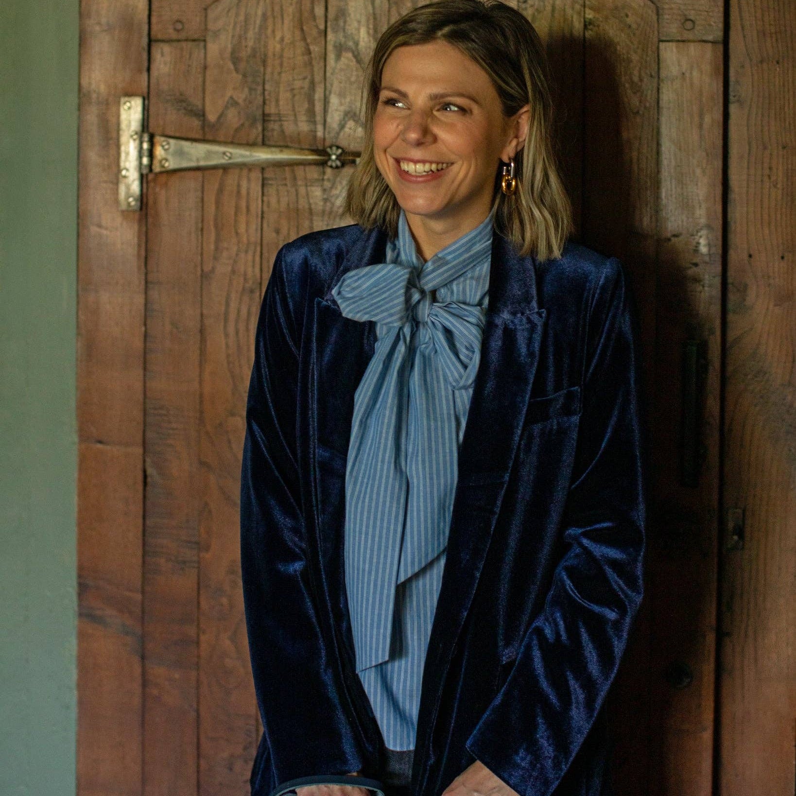 A woman with blonde hair stands smiling in front of a rustic wooden door, wearing a dark navy blue velvet blazer over a light blue and white striped blouse with a large bow detail.