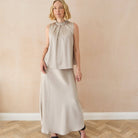A woman modeling a sleeveless high-neck two-piece dress in a light beige or oatmeal color, featuring a cropped top and a long skirt, standing against a beige wall with a herringbone wood floor.