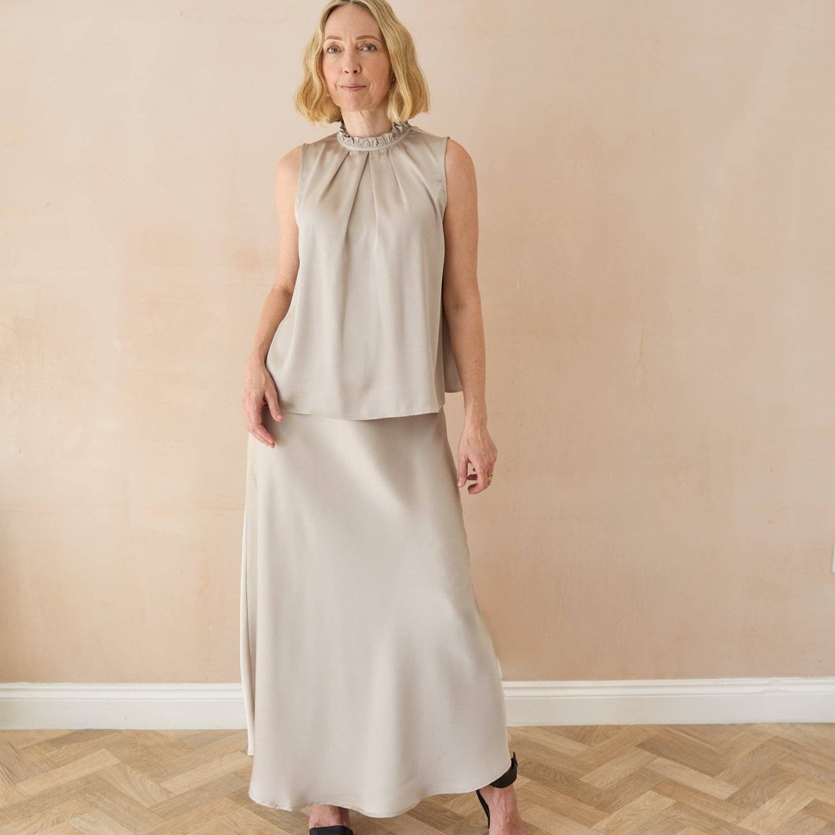 A woman modeling a sleeveless high-neck two-piece dress in a light beige or oatmeal color, featuring a cropped top and a long skirt, standing against a beige wall with a herringbone wood floor.