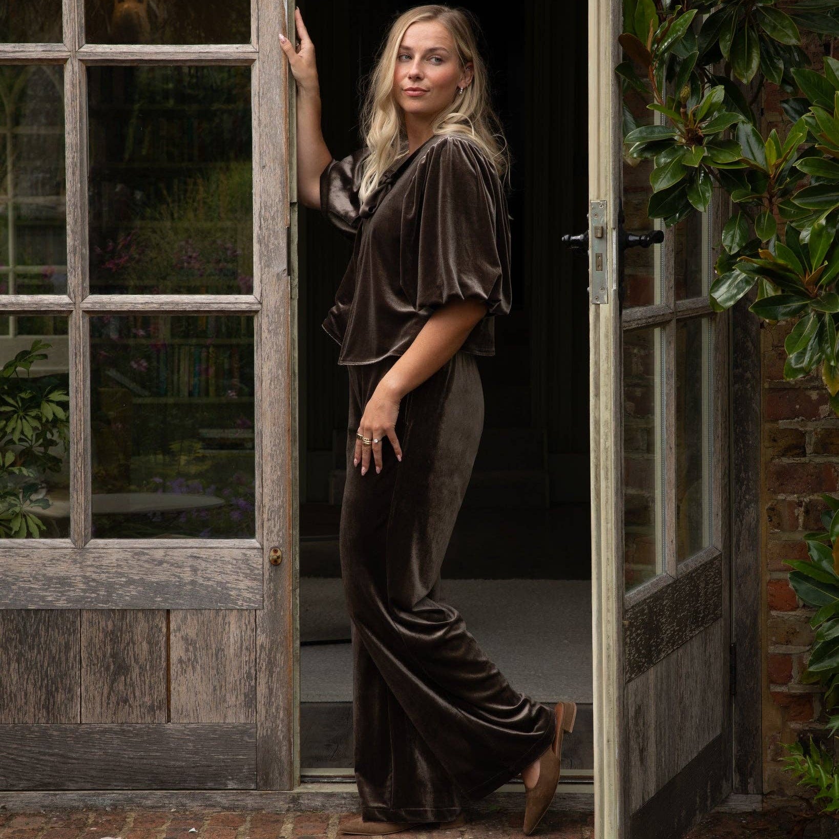 A woman stands in an open rustic wooden doorway wearing a dark brown velvet two-piece outfit consisting of a short-sleeve top and matching wide-leg trousers, with brown shoes.