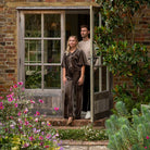 A woman in a dark brown velvet jacket & trousers stands in the doorway of a rustic brick house, with a man in a beige t-shirt and dark pants behind her. The scene is framed by lush garden foliage, pink flowers, and a stone path.