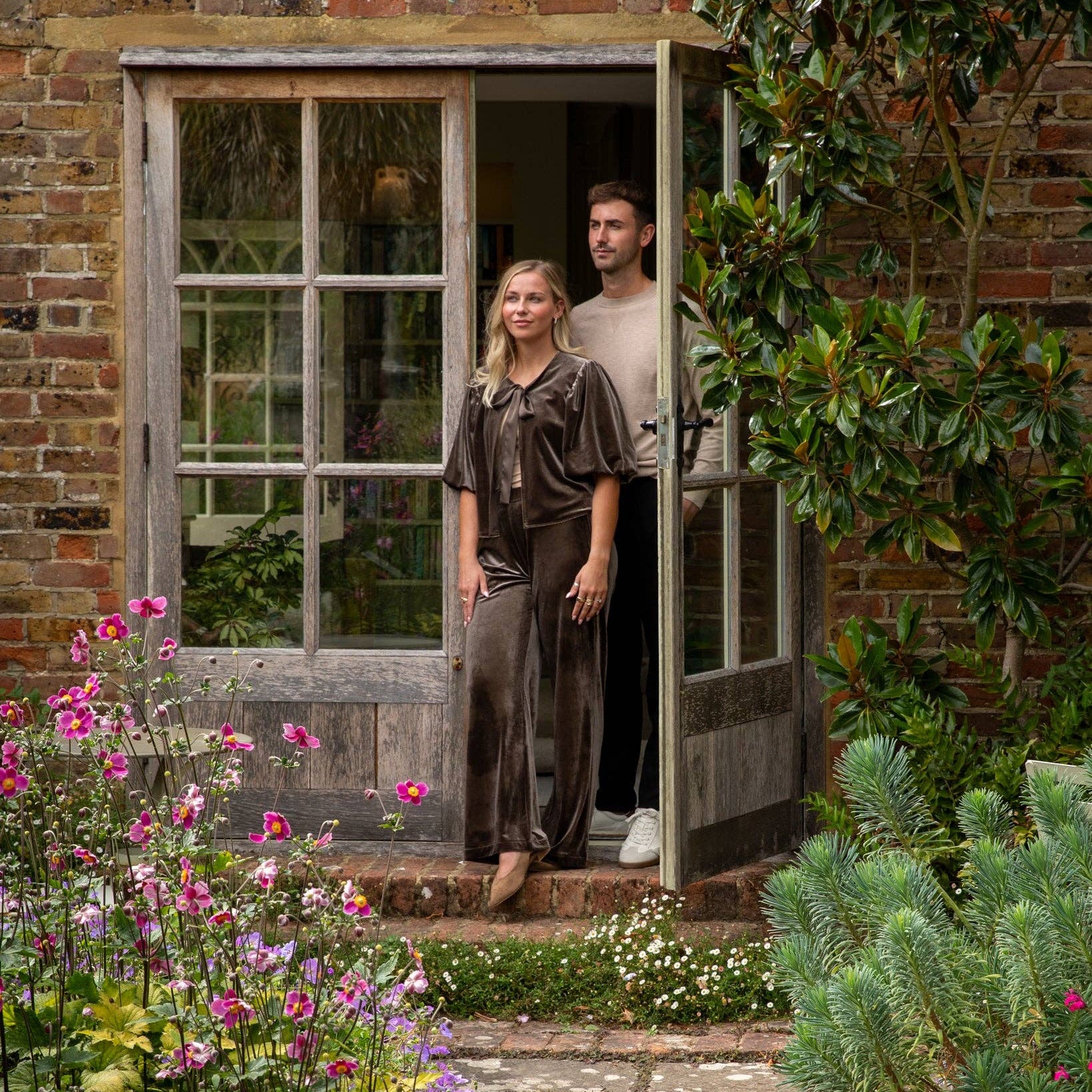 A woman in a dark brown velvet jacket & trousers stands in the doorway of a rustic brick house, with a man in a beige t-shirt and dark pants behind her. The scene is framed by lush garden foliage, pink flowers, and a stone path.